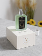A Voreza London Obsidian Perfume bottle with a silver spray cap and black label, displayed on top of its white branded box. The bottle is a pale mint green, set on a gray stone surface with a blurred background of dried grass and yellow flowers.